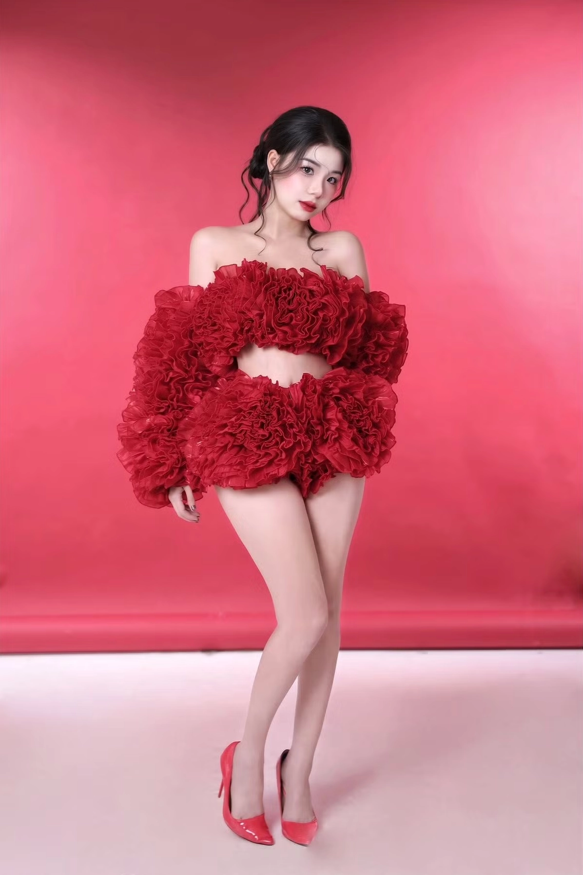 Linh hồn của bộ ảnh của hot girl Trà Hạ Ly chính là bộ trang phục màu đỏ rực rỡ. Đây không phải là một chiếc váy thông thường mà là một set đồ 2 mảnh, bao gồm áo crop top trễ vai và quần short, được tạo nên từ vô số lớp vải voan xếp nếp bồng bềnh. Thiết kế độc đáo này trông tựa như một đóa hồng nhung khổng lồ hay một đám mây bồng bềnh, vừa che vừa hở.